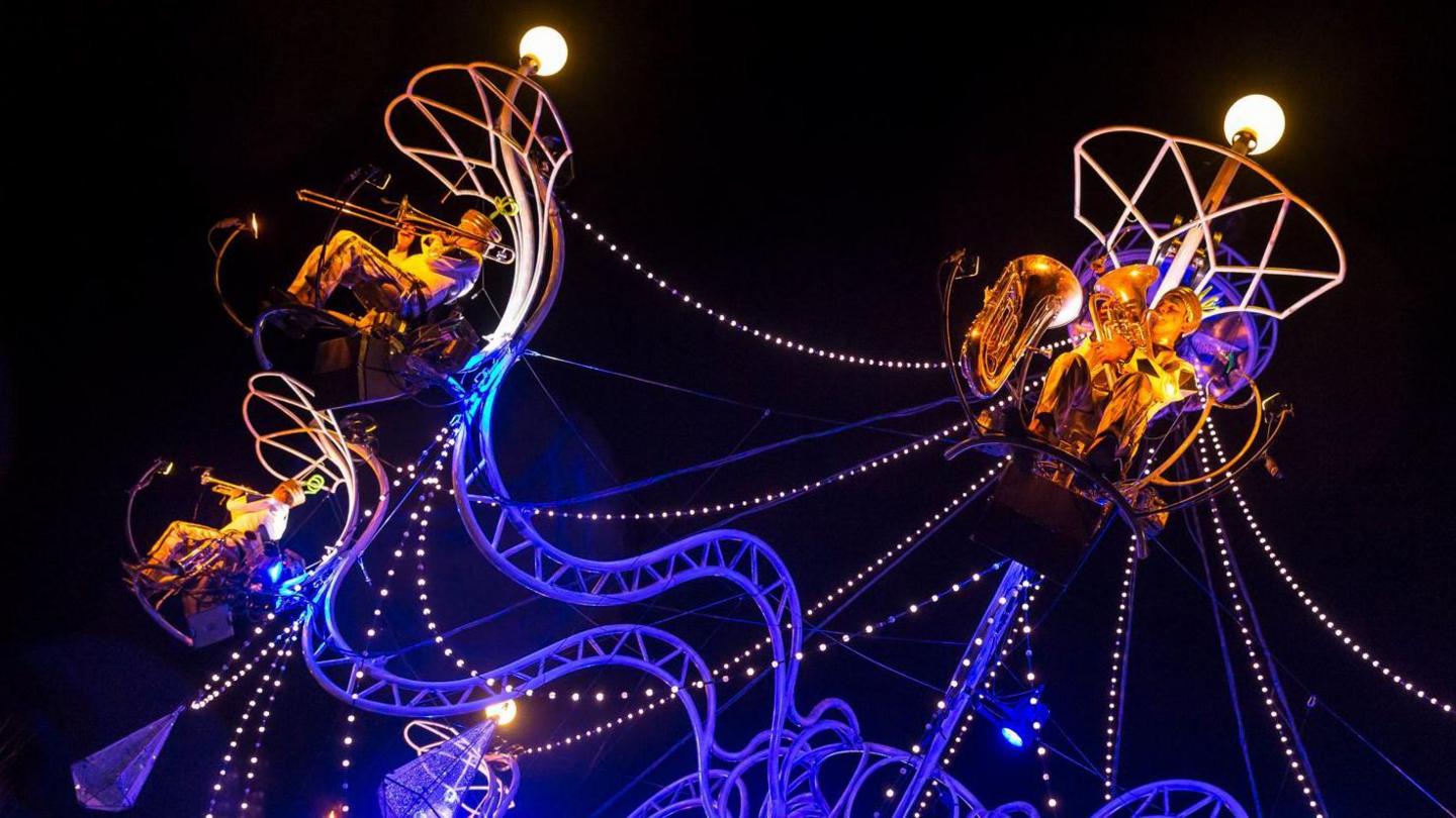 Musicians are performing on a giant chandelier. They are lit up in yellow under lamps on the large chandelier structure during a performance at night.