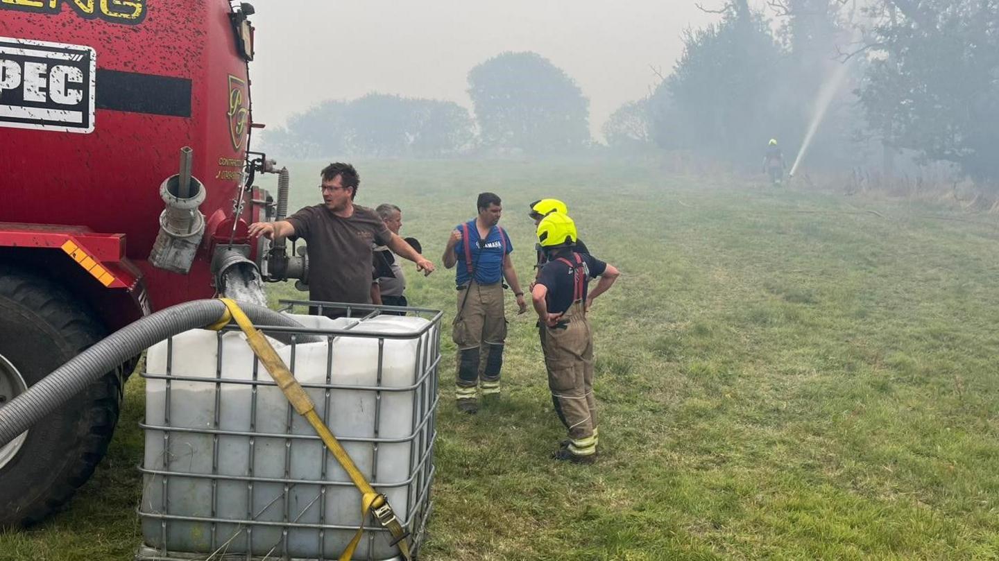 A man is filling a large container with water from a tanker in a field with a fire officer nearby and smoke pouring from a fire in the distance