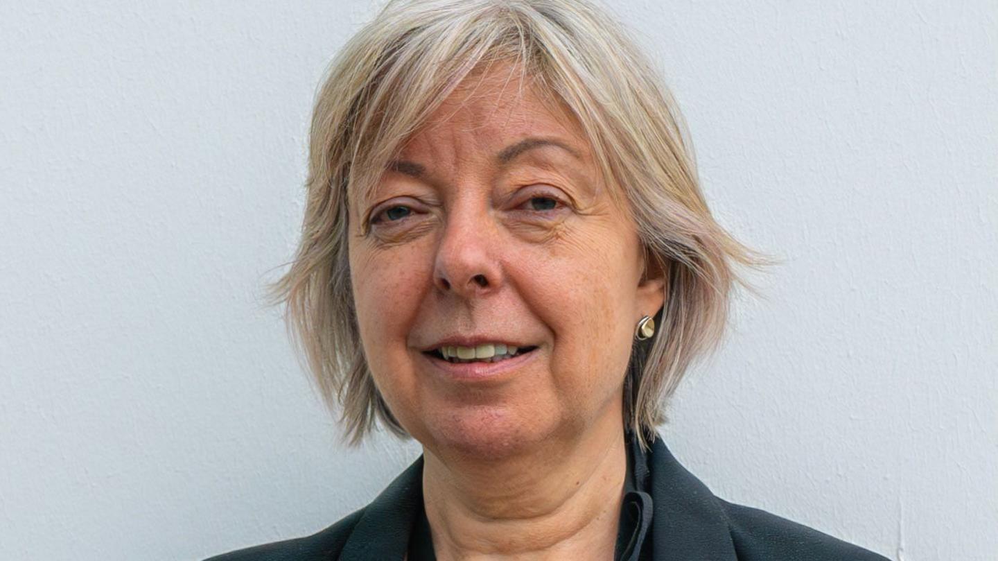 Karen Ramsay, a woman with short blonde hair. She is wearing a dark blazer and standing against a white background while looking at the camera.