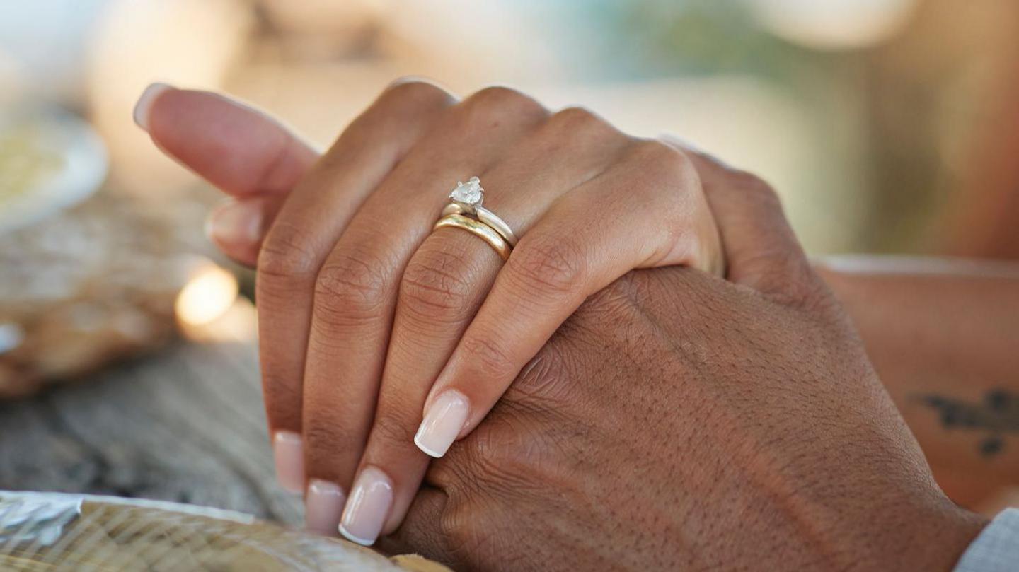 A woman's hand, with wedding and engagement rings on the ring finger clasps a man's hand.