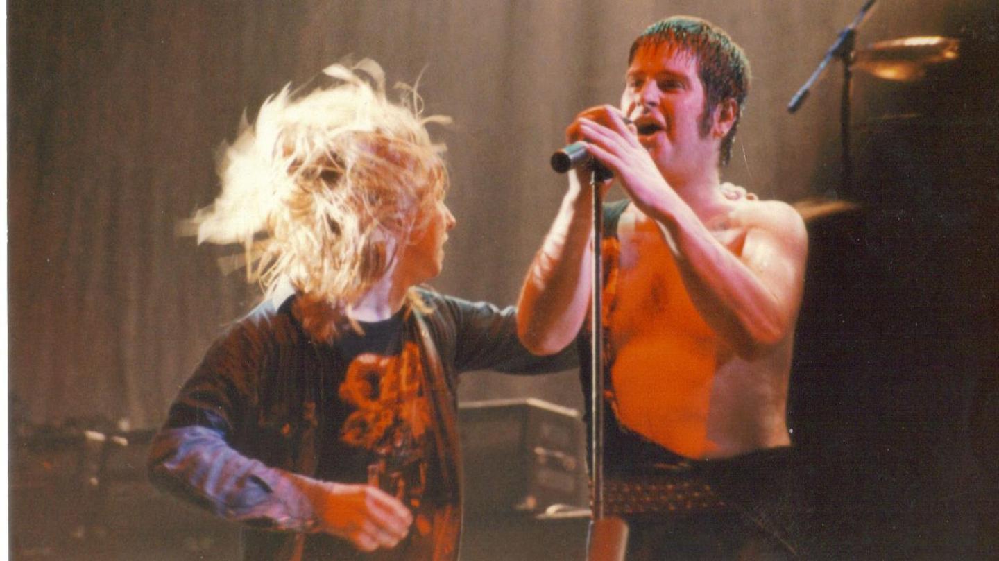 Des Rumney with long blonde hair, goes to hug Ozzy Osbourne on stage. Ozzy is shirtless as he is singing into a microphone.