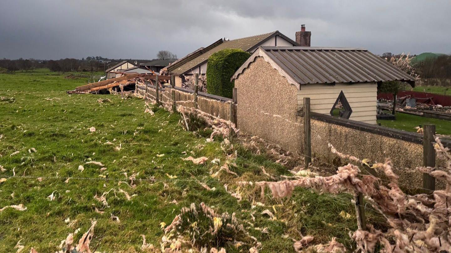 The insulation of a roof is strewn across a field and a house is damaged