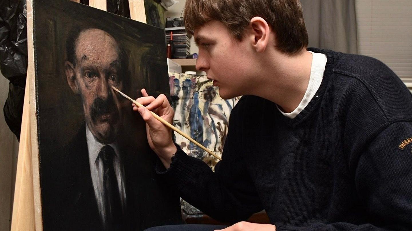 Connor Draycott. who has short light brown hair and is wearing a navy jumper, makes finishing touches to his portrait of James Finlayson with a wooden paintbrush.