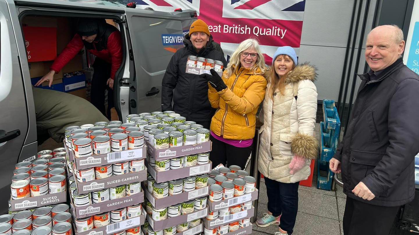 People are pictured in front of a van next to stacks of tins. There is 4 people posing for a picture with one person in the background in the van.