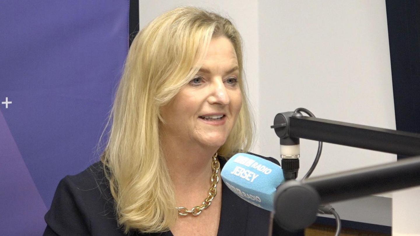 speaking into a blue microphone which has the words 'BBC RADIO JERSEY'. She has blonde hair and is wearing a gold chain necklace and a black top.