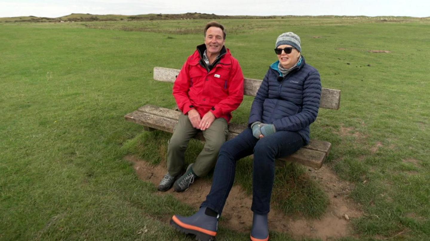 David Brain sits on a bench wearing a red jacket and Liz Brady has a grey woolly hat and dark glasses, a navy padded jacket, gloves and boots and is wrapped up against the cold.
They are sitting on a bench looking out across the countryside.