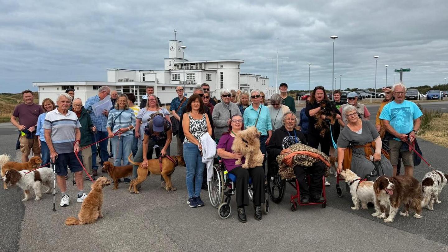 About 30 people, many with dogs, gather on Birkdale Common to protest plans to allow the R&A to take over a section of the public land ahead of the 2026 Open golf championships there. Three people in wheelchairs sit in the foreground, in front of the standing protesters.
