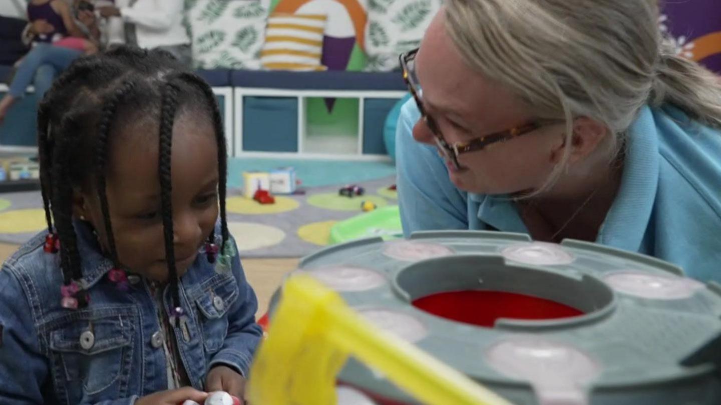 A woman wearing glasses on the right who works for the parenting network, she is playing with a young girl on the floor while parents can be seen in the background talking