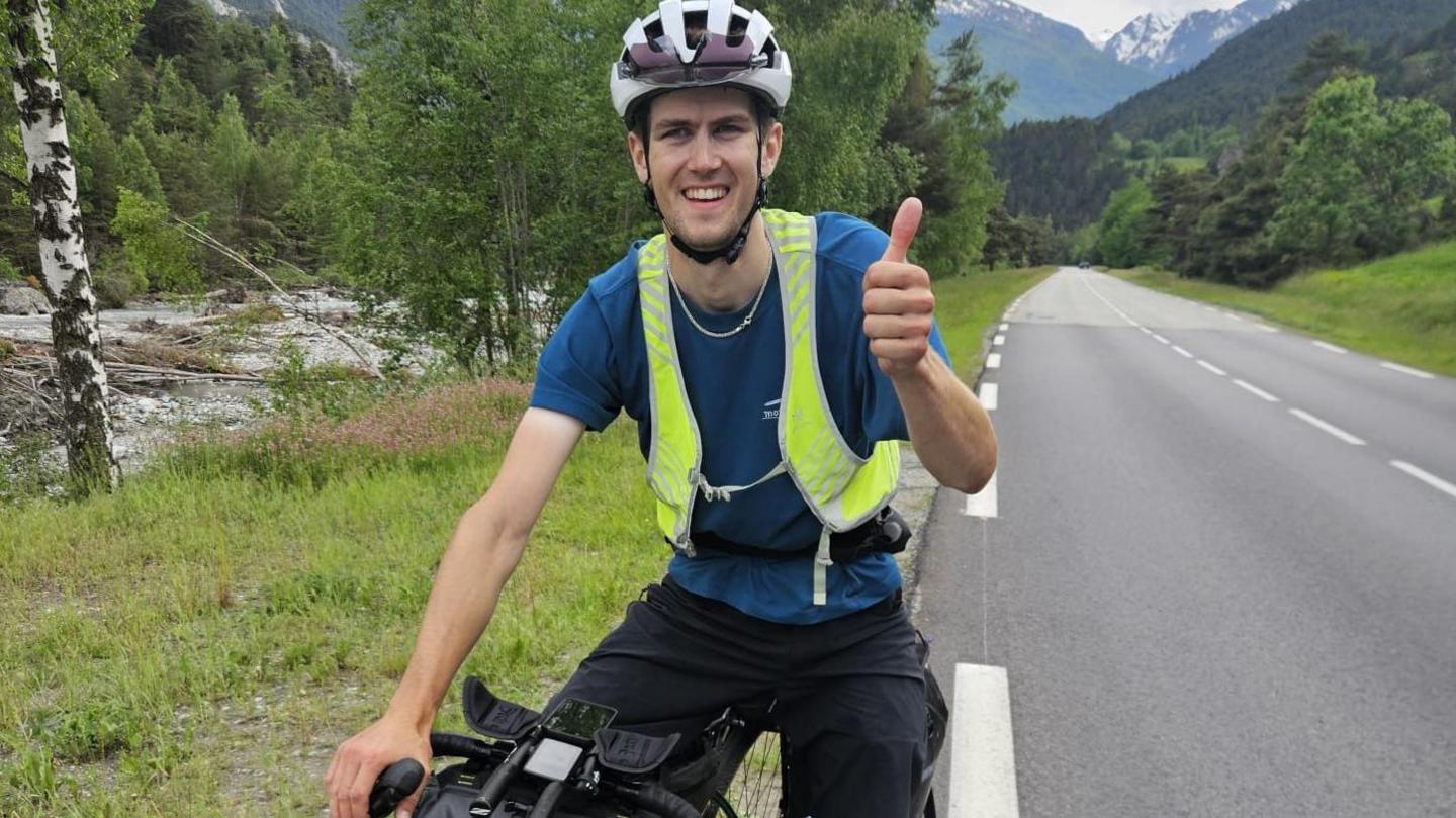 Finlay Tusting is on a bicycle and wearing a bike helmet, high vis jacket, blue T0-shirt and black trousers. He is on the side of an empty road with snow-topped mountains behind him and giving a thumbs up to the camera.