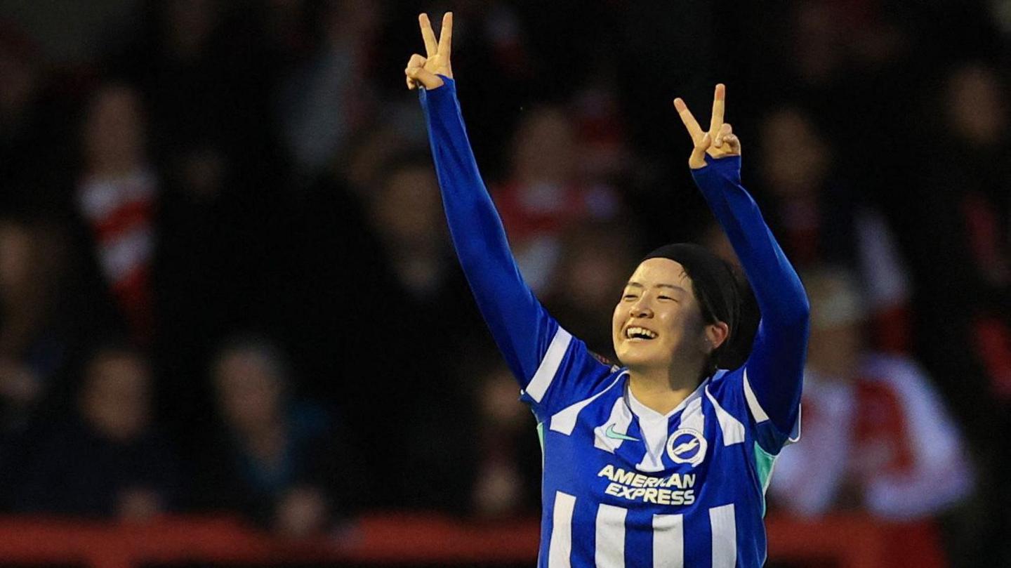 Fuka Tsunoda, wearing a blue and white Brighton shirt with long blue sleeves, smiles and raises her arms above her head, with her index and middle fingers making 'v' shapes, celebrates