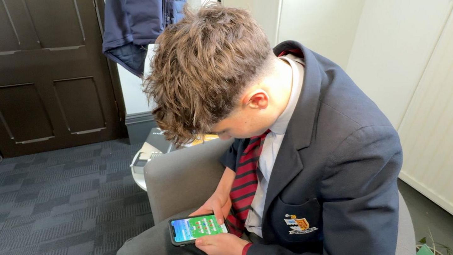 Student in suit and tie sat in a chair. he's holding his phone and has the Fantasy Football app open.