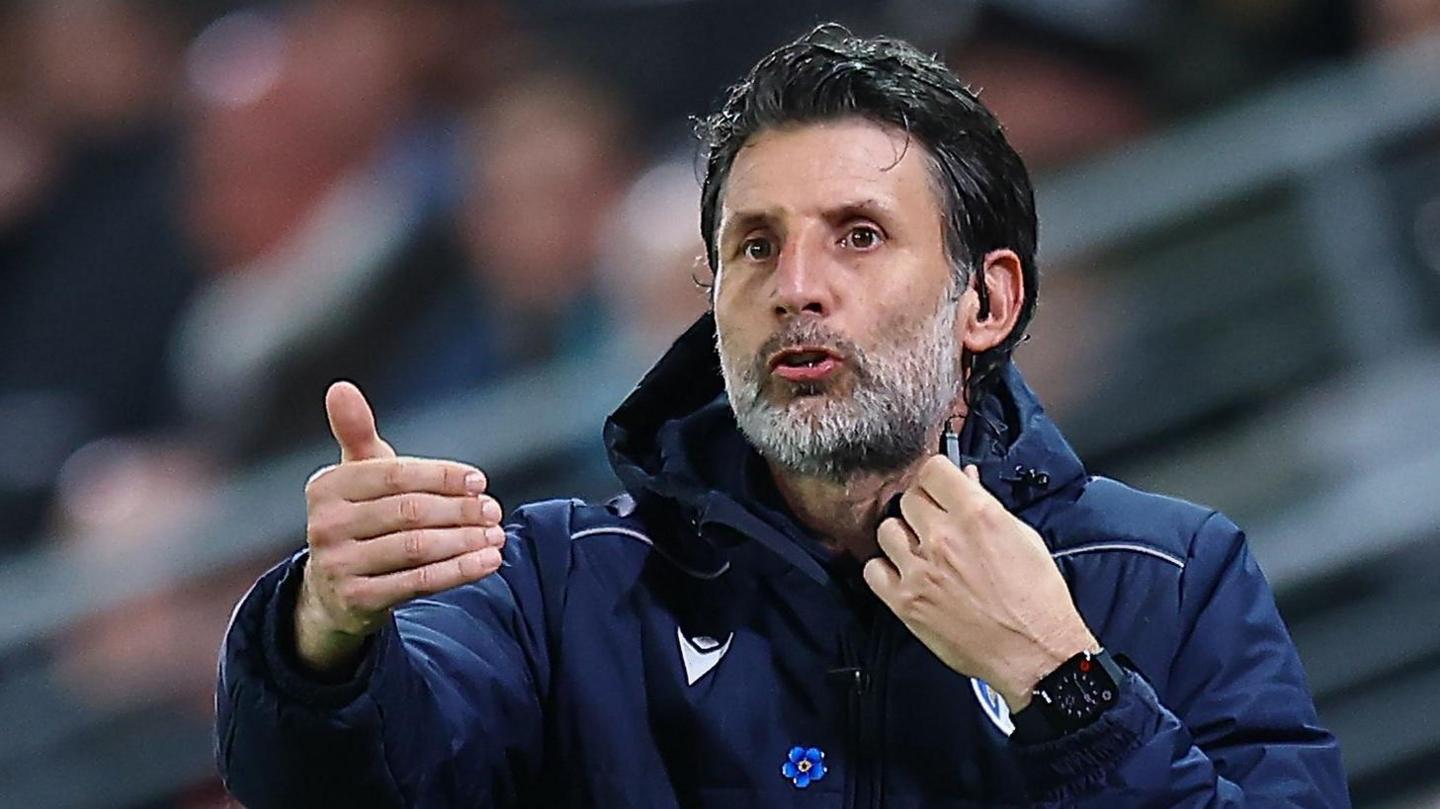 Danny Cowley, in a blue training jacket, issues instructions from the sidelines at MK Dons to his Colchester side