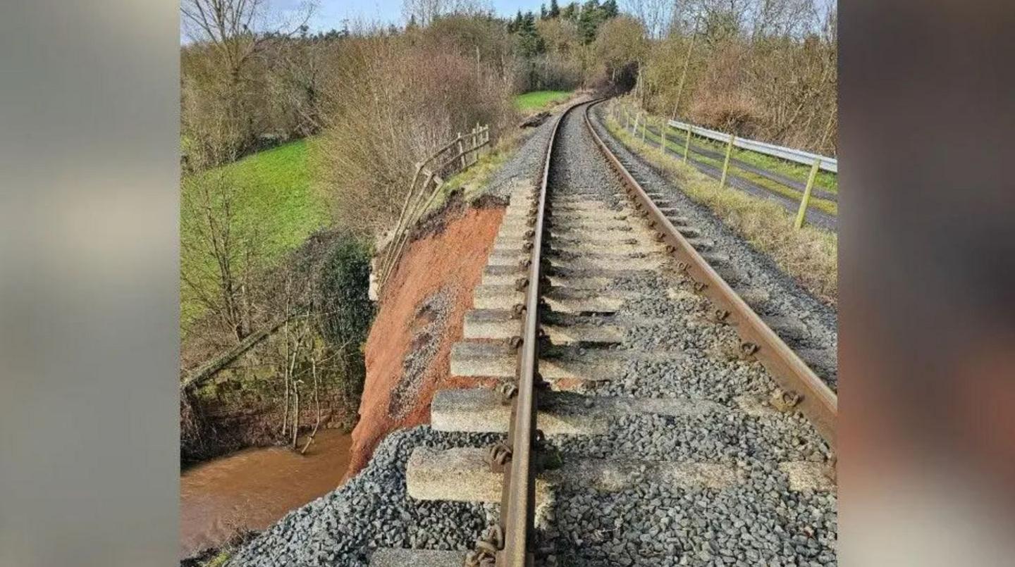 A railway line running through an area of grass and trees with the soil on the left hand side exposed and a section of the ground beneath it missing