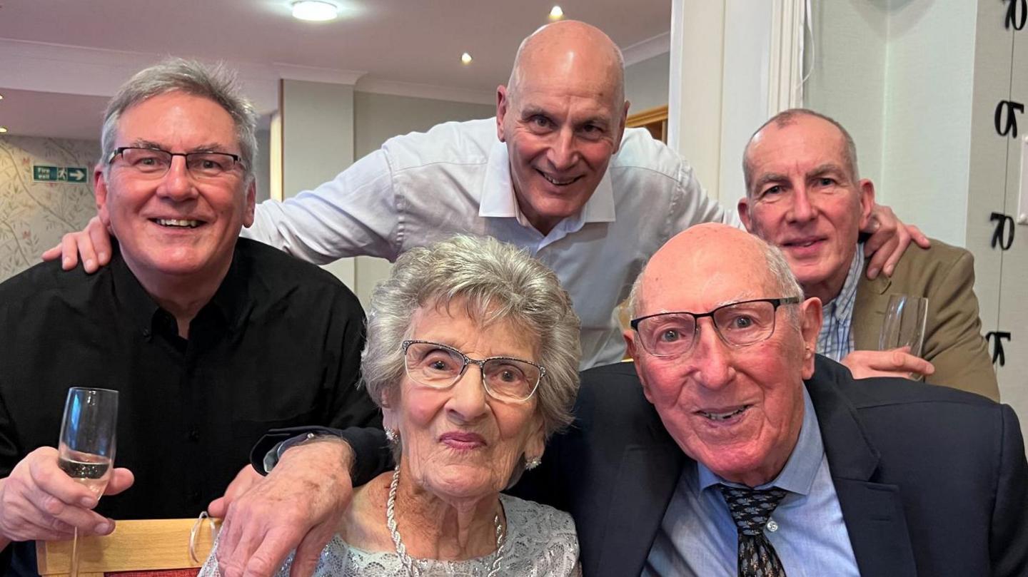 Mr and Mrs Ashpool are joined by their sons David, Tony and Mick at the anniversary party. The three men are wearing shirts and smiling while standing behind their parents.