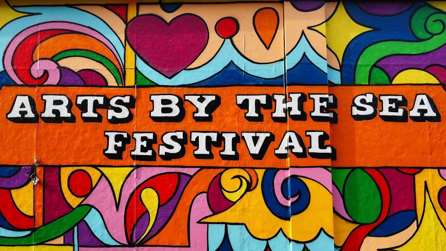 A multi-coloured spray painted mural on a wall which says "Arts by the sea festival"