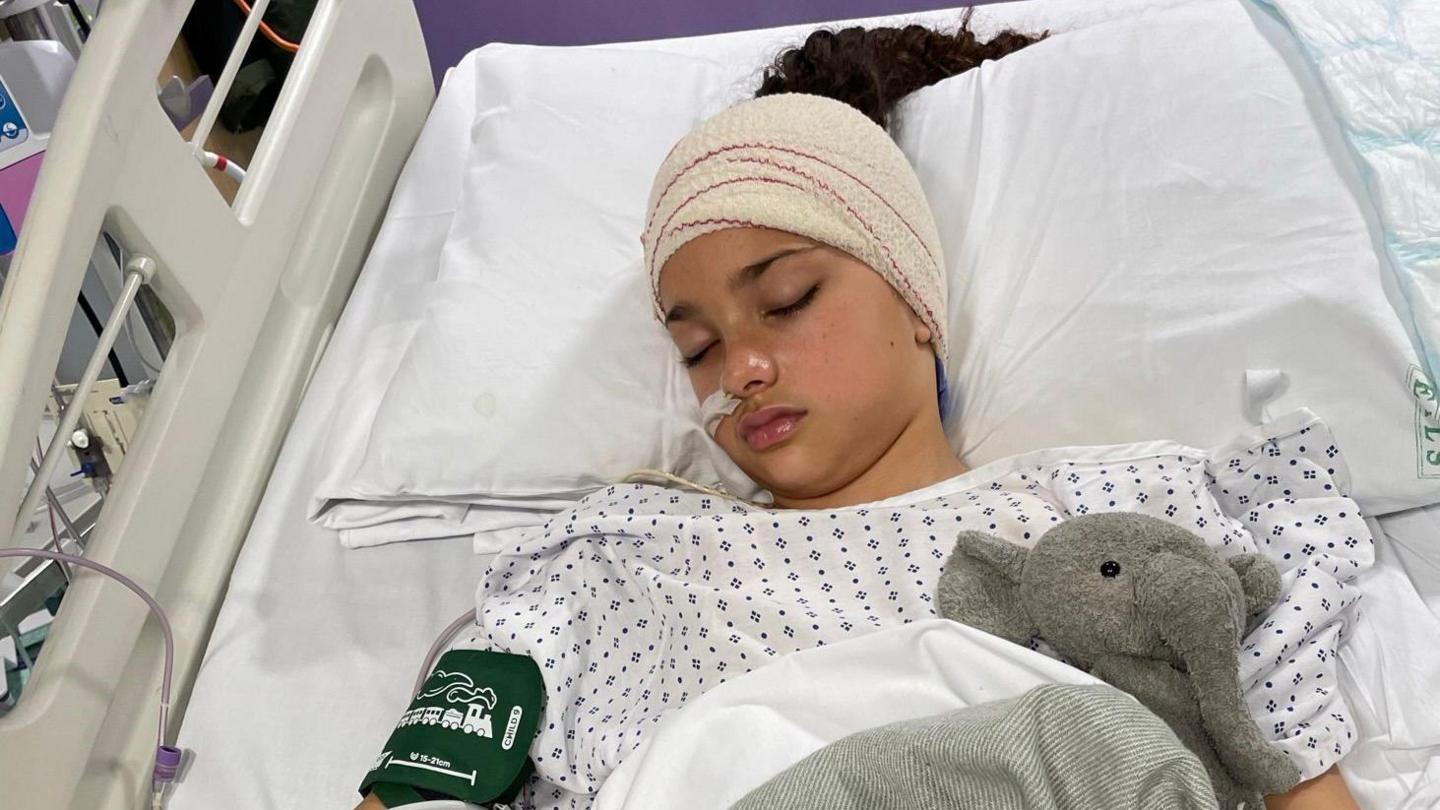 Amelie lying in a hospital bed wearing a white hospital gown. She is unconscious and has her head wrapped in bandages and a pressure cuff on her upper arm. She is also snuggled up with a teddy elephant under her blanket.