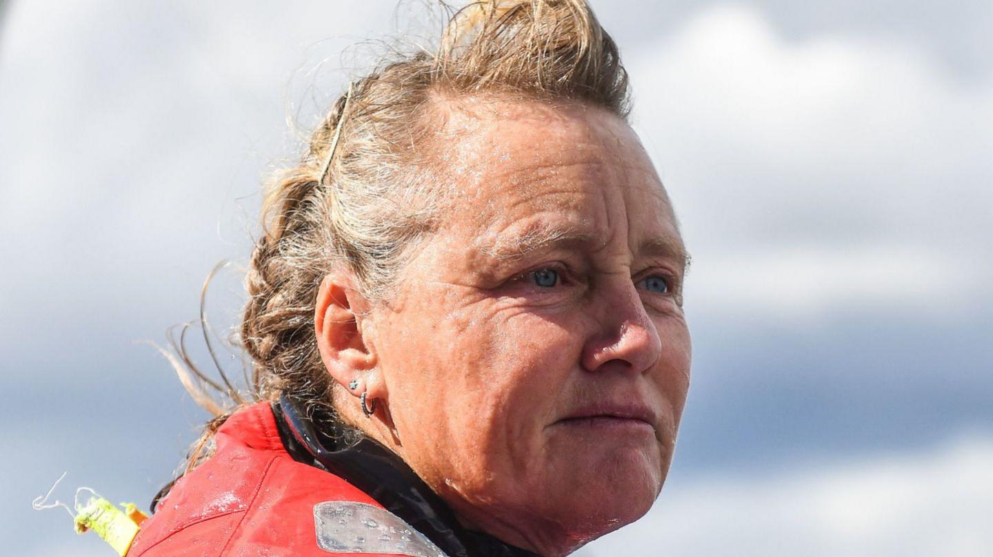 Pip Hare looks over the edge of her yacht toward the sea as she fastens a rope around a metal pole. Her blonde and grey hair is windswept and she is wearing a large red waterproof coat