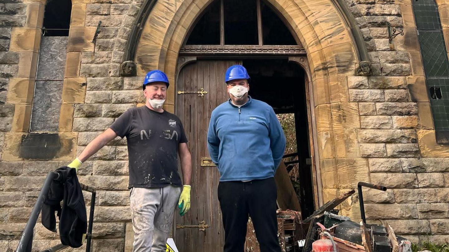 Paul Taylor (left) standing outside the church entrance. Large wooden doors with brass hinges stand in an arched doorway. He is standing next to another man who is helping with the clearing work. A pile of rubbish, including an old fire extinguisher, is on the floor next to them.