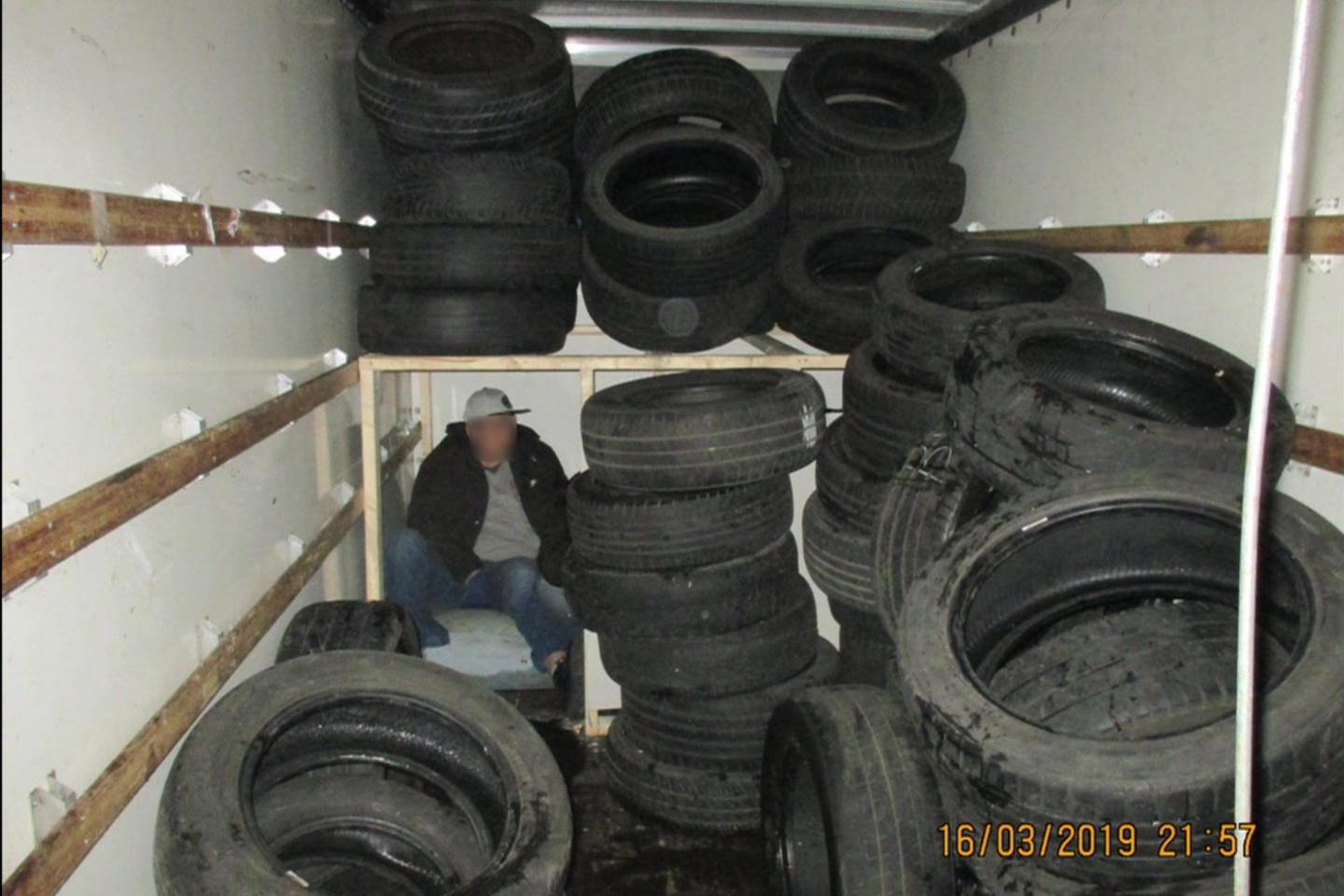 A wider view of the vehicle's interior shows one man sitting inside a makeshift compartment among numerous stacks of car tyres. The wooden panels of the compartment are clearly visible, and the space appears dimly lit. One man’s face is blurred for anonymity. The timestamp "16/03/2019 21:57" is visible in the bottom corner of the images.