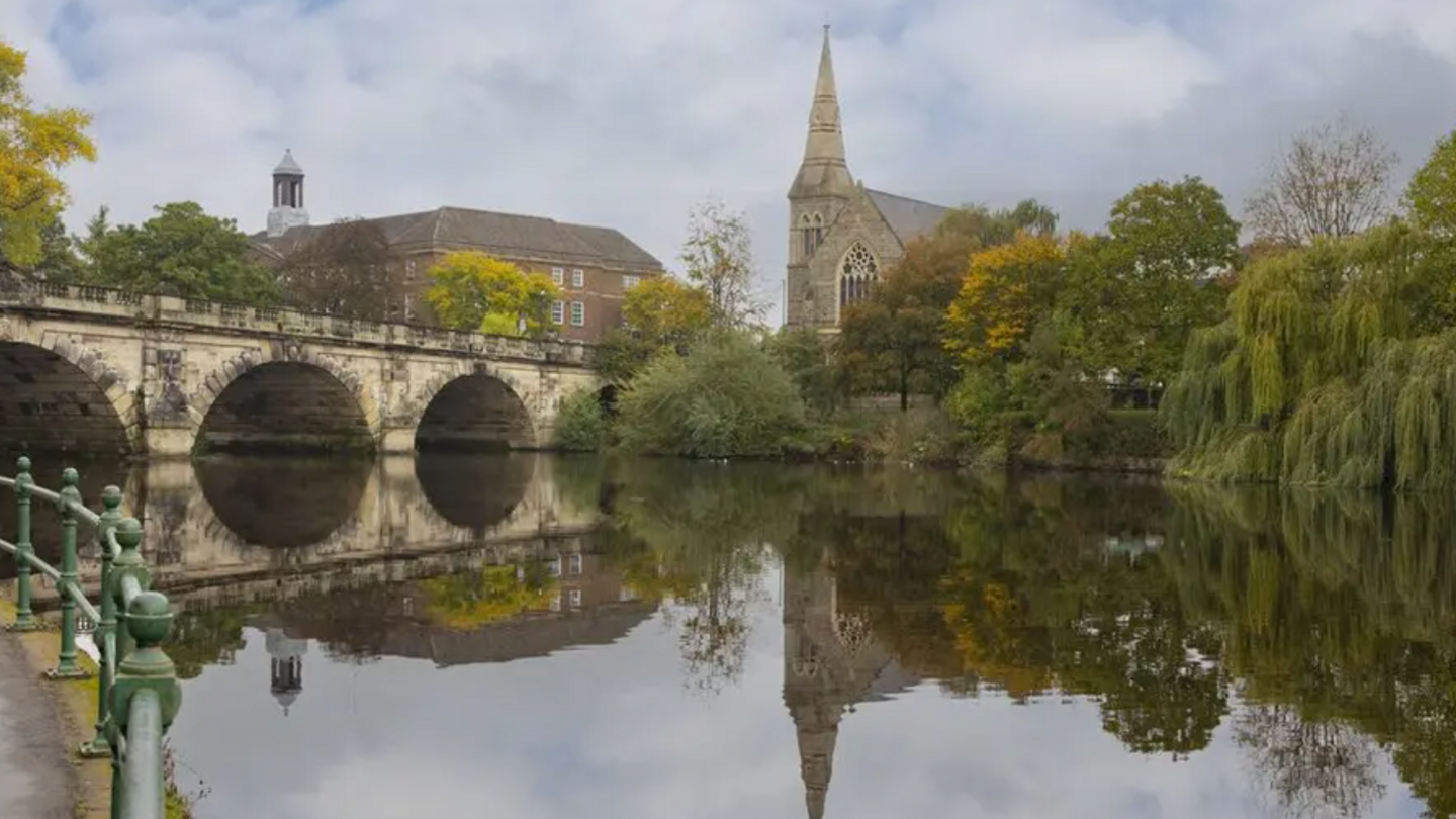 The River Severn in Shrewsbury, with a bridge, buildings including a church and trees in the distance that can be seen reflected in the water, and a walkway with railings on the left.