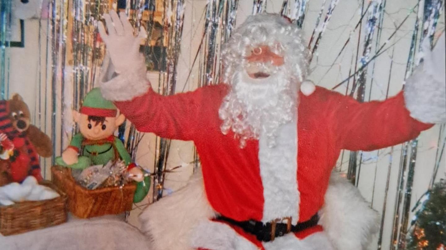 Mick playing Father Christmas in a grotto. He is wearing a Santa suit and has his arms up posing for the camera. Two baskets of toys sit behind him. 