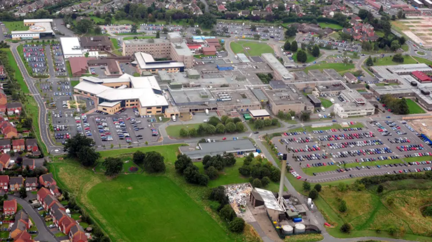 An aerial view of a hospital site, with a number of large buildings visible, surrounded by a car park and green fields, with a street with red brick houses to the left hand side