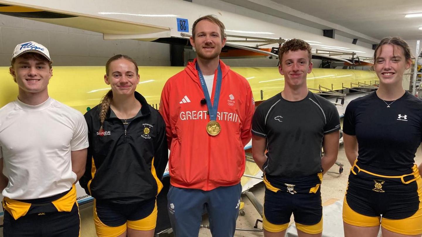 James Rudkin stands besides four members of Northampton Rowing Club. James is wearing a red Team GB tracksuit top and his Olympic gold medal. The others are standing either side of him, with their hands behind their back and smiling at the camera. In the background you can see rowing boats.  