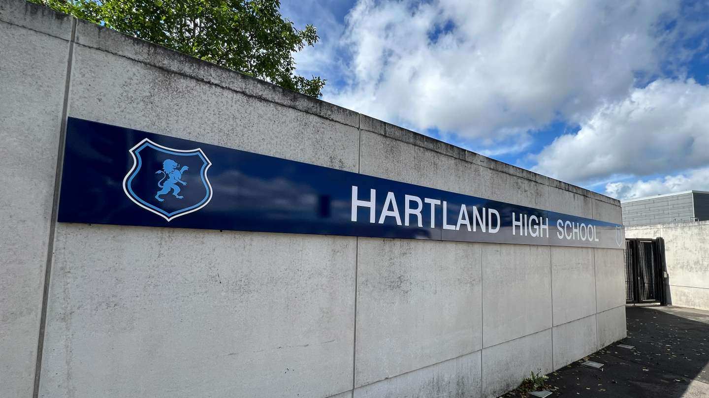A general view of a sign that says Hartland High School on it with a new logo with the outline of a blue lion in a crest on the far left side of it. It is attached to concrete at the school's entrance.