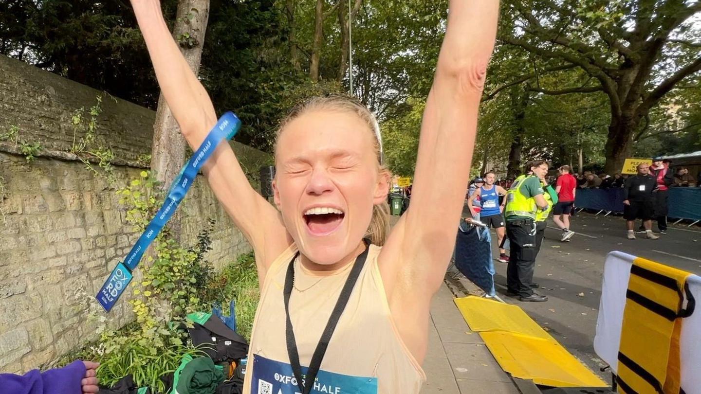 Harriet freeman, wearing a beige vest, has her arms a loft in the air. In one hand is a medal.