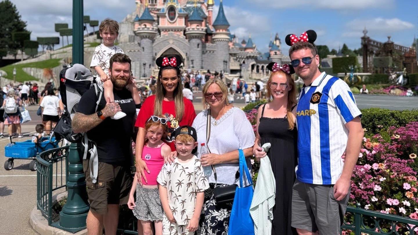 A group of people are standing in-front of a Disney castle wearing Mickey Mouse ears. Alison is in the middle. There are three young children, Alison’s grandchildren