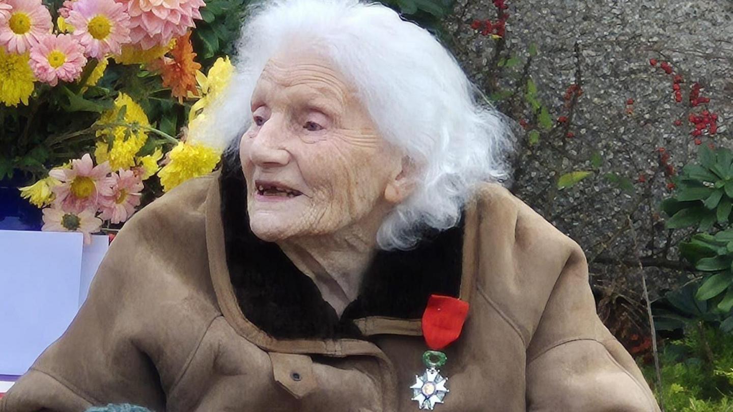An elderly woman with grey hair in a brown coat has a medal with a red ribbon pinned to her lapel