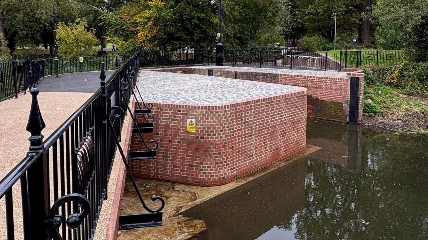 The newly-repaired bridge. It has black railings and runs over a fairly narrow river. Its side consists of fresh red bricks and the surface is made of grey bricks.