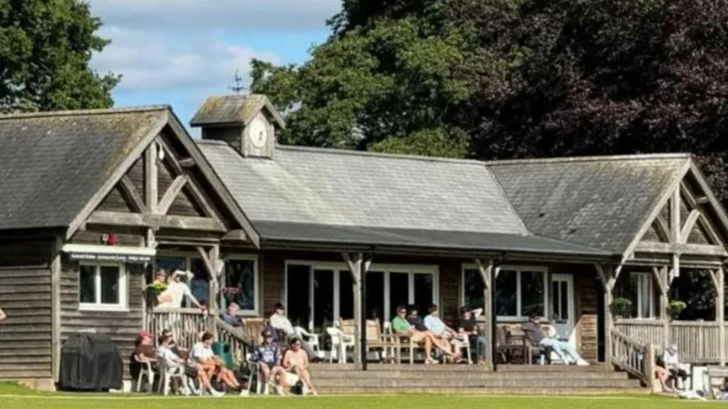 Heathcoat Cricket Club's wooden pavilion before the fire. About 15 people are sitting outside the single-storey structure, which had a small clock tower and partial glass frontage