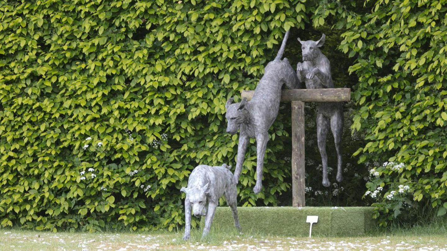 A sculpture depicts three dogs emerging from shrubs and bushes