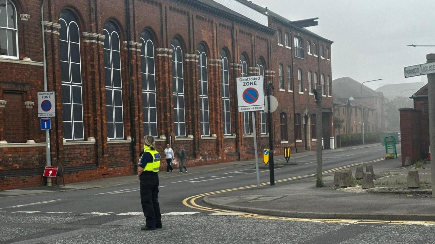 A police officer wearing black trousers and yellow high visibility vest stands in the centre of a road. A large brick building is visible in the background which is covered in hazy grey smoke.