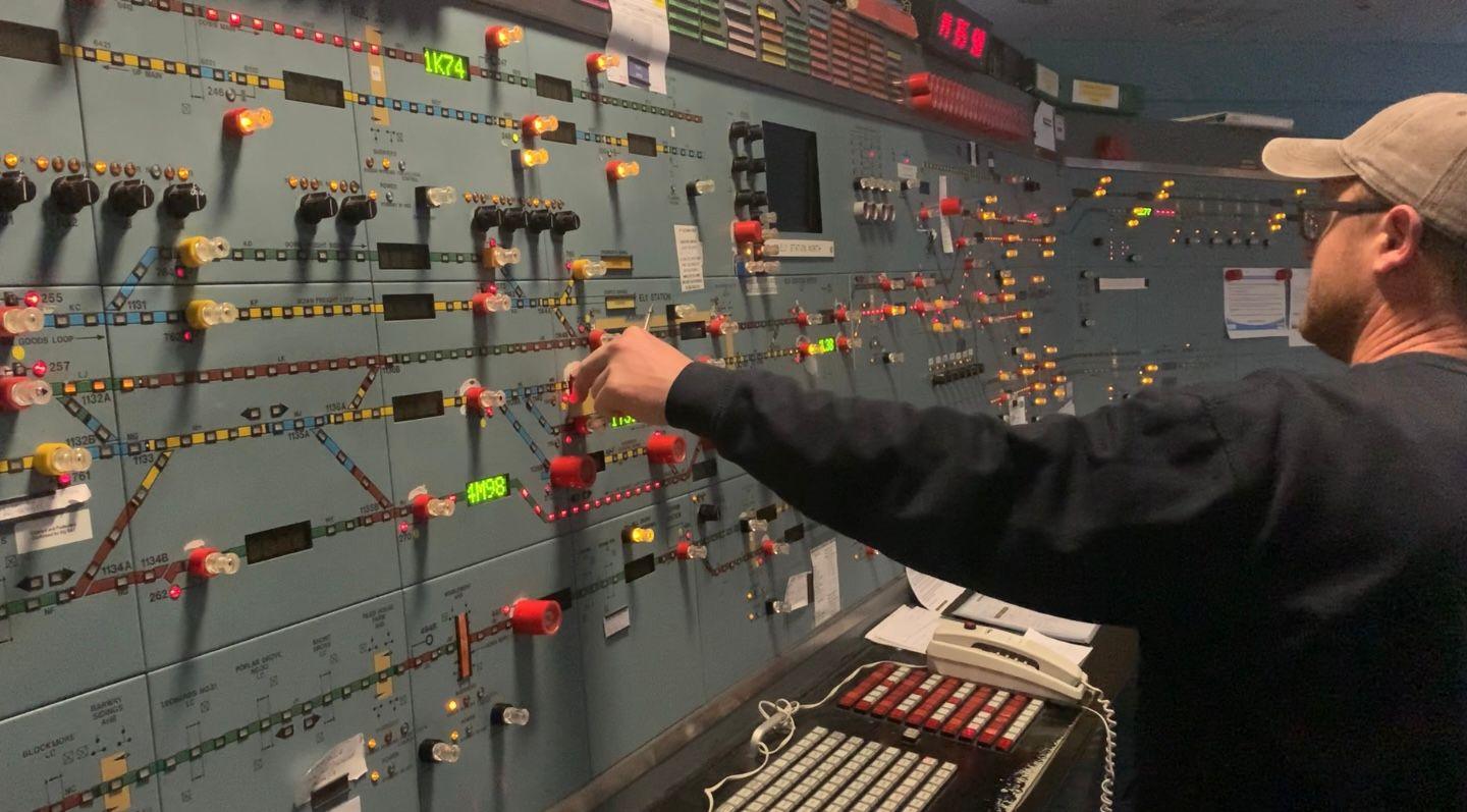 The inside of a signalling centre, with grey/blue panels. There are lots of knobs and buttons. some lit up, and a rail map on the walls. A man wearing a beige baseball cap and black sweater stands with his back to the camera and twiddles a red knob. 