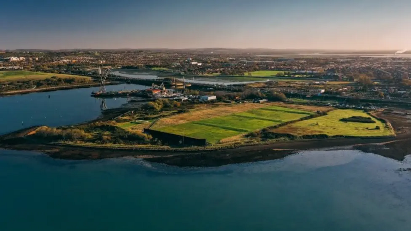 An aerial view of the section of the peninsula Tipner West where plans are set to take place. There is a section of the island which is covered in grass field, lines by trees and surrounded by water.