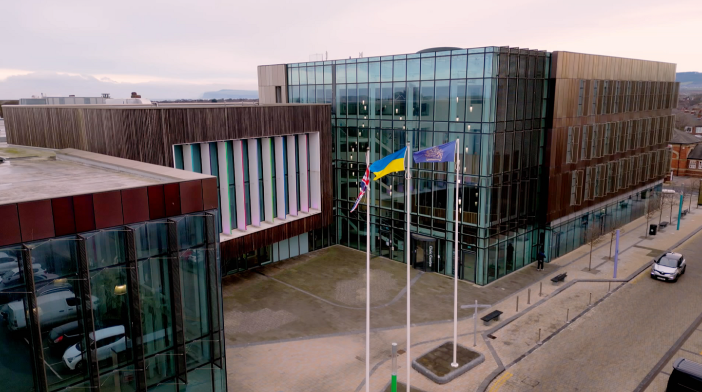 Redcar and Cleveland Council's headquarters photographed from high up. There are three, interlinked square brown buildings. They are clad with the two on either side containing large glass frontages. The middle building, which sits back slightly from the other two, has a large area of wood-effect cladding with windows in the front separated by long, thin white pillars. In front stand three large flagpoles flying the flags of, left to right, the United Kingdom, Ukraine and the council's crest in gold on a blue background.