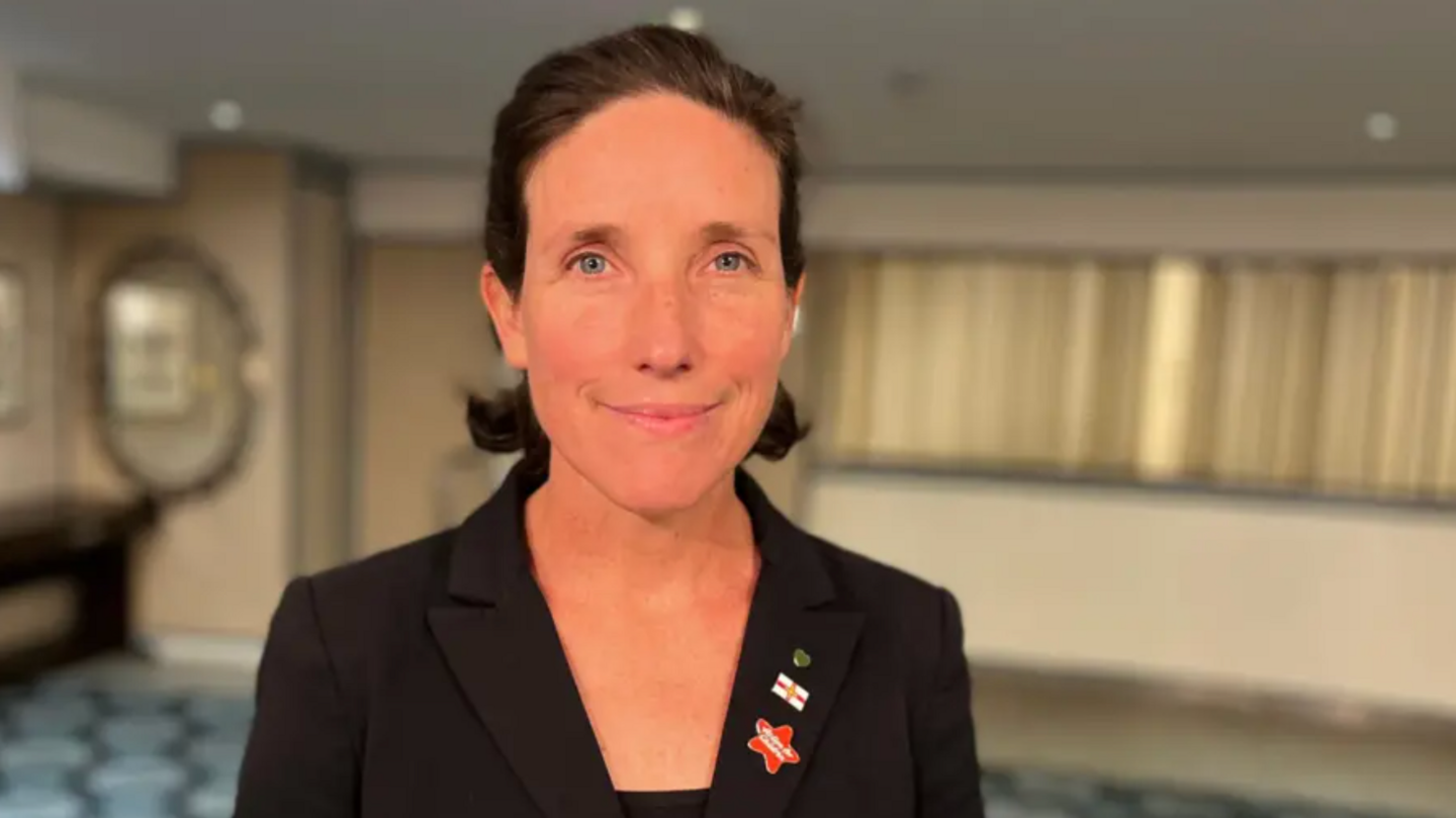 Deputy Lindsay de Sausmarez stands looking at the camera wearing a black jacket with broaches on the collar. She smiles with a blurred room behind her.