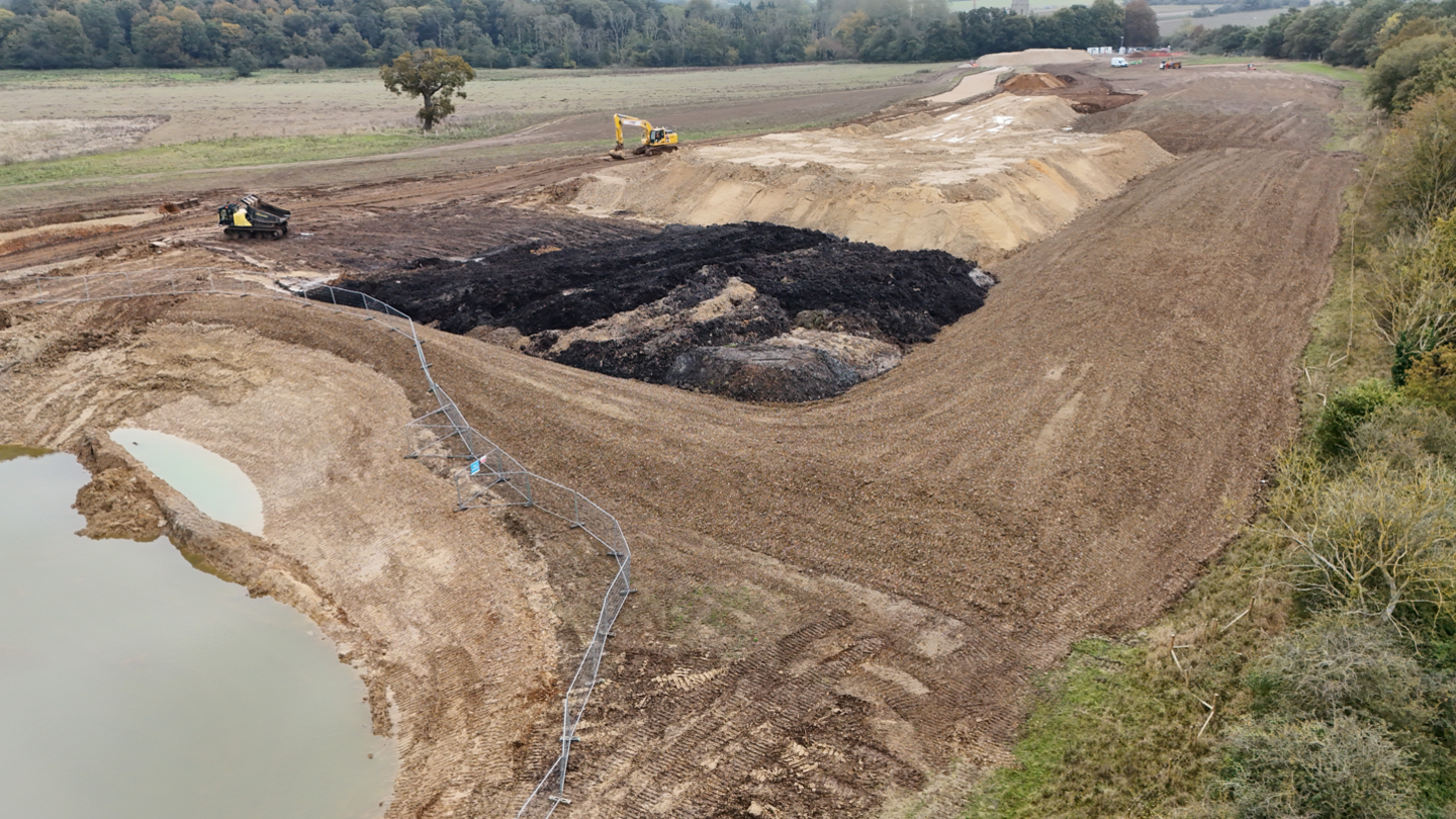 A part of a field has been dug up, with the middle section of the picture showing a big pile of dirt. At the bottom left of the picture is a big hole filled with water, with a silver wired fence around it. 