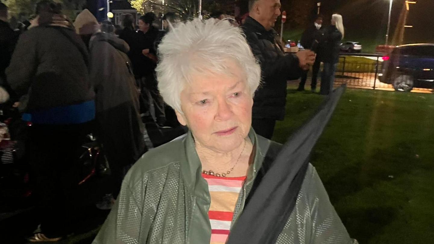 An older woman, with short grey hair, is standing outside on a city street. In the background there is a crowd of people, all wearing coats and hats to protect form the rain and cold. There is a road too, with a car going past. The woman is holding a black flag, in tribute to the Bloody Sunday dead, and wearing a green coat.