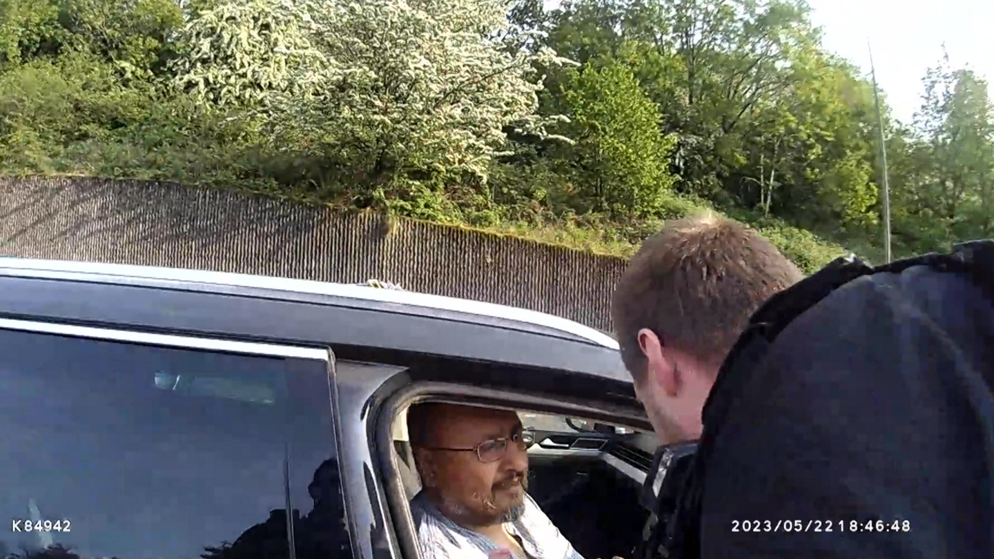 Still image of police bodycam footage of a man getting arrested in a dark coloured taxi by a police officer wearing black on a busy road.