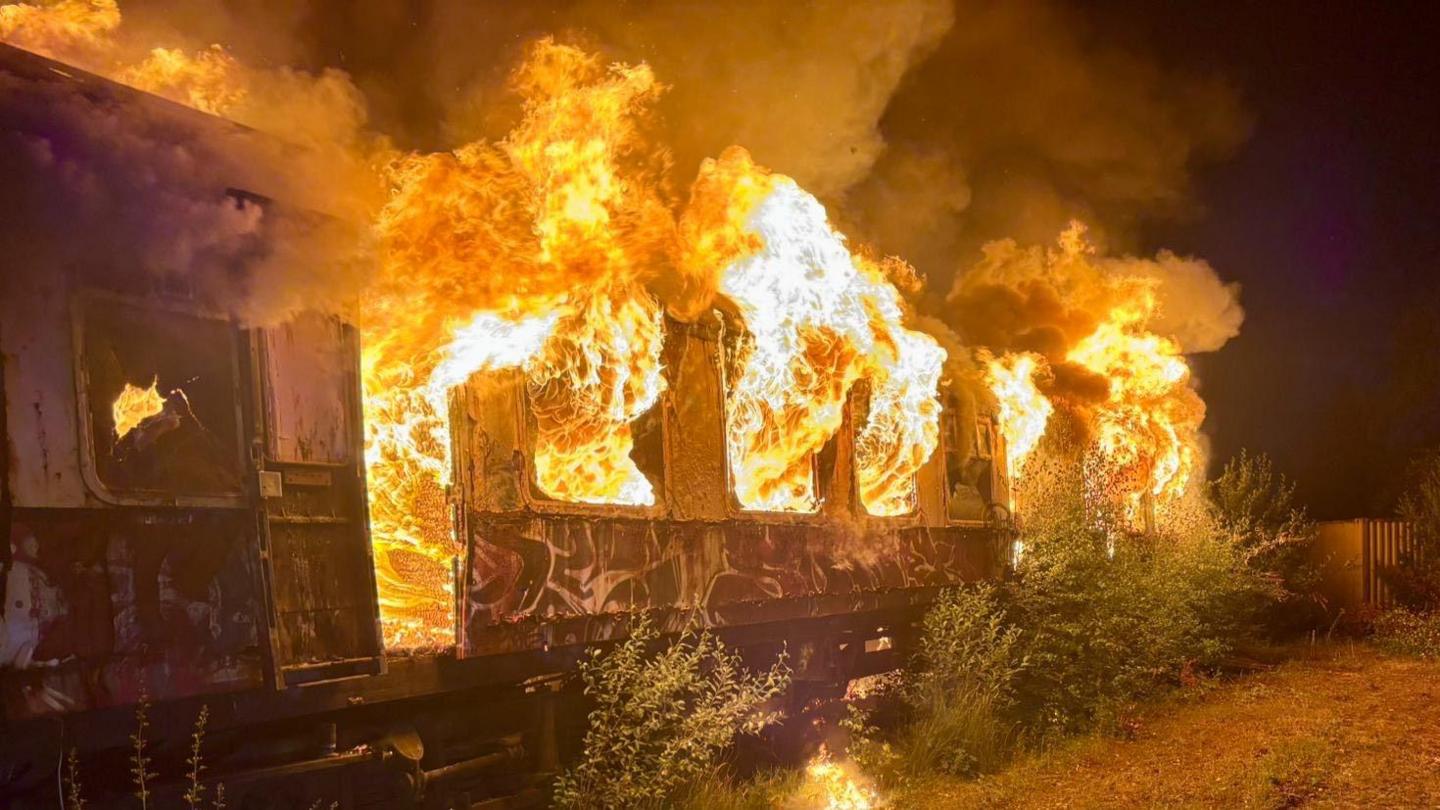 A train carriage with flames bursting out of the windows. There are bushes growing near the carriage, and fencing in the distance.