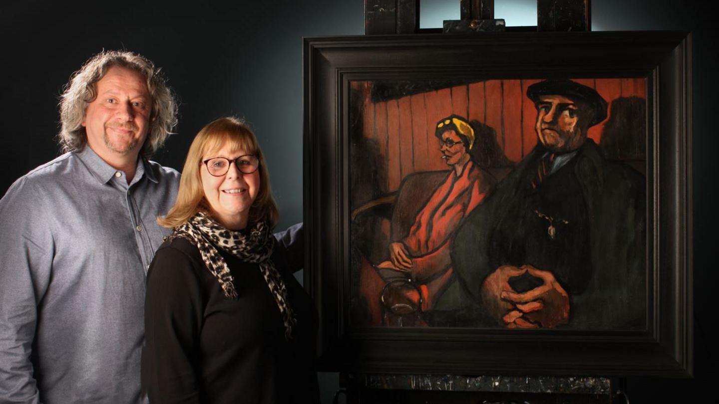 Richard Hindle and Corinne Aspel standing next to one of Tom McGuinness' paintings. Mr Hindle is wearing a grey shirt. Ms Aspel is wearing a black shirt and glasses and a leopard print scarf. The painting depicts a man and woman sitting in chair. The man is smartly dressed and is wearing a flat cap. The woman has a yellow headband and a red coat and is smoking.