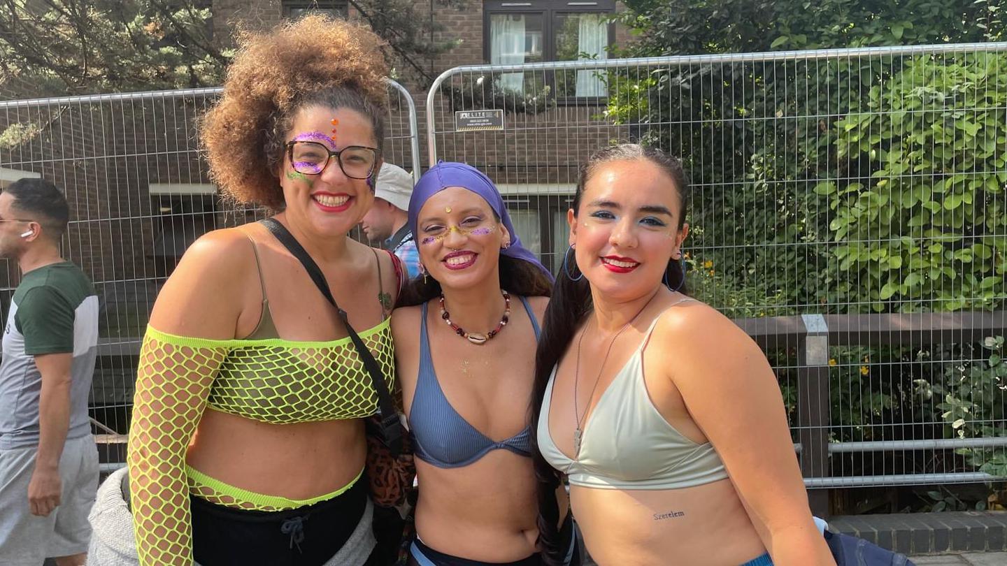 Two women at the Notting Hill Carnival are dressed in vibrant, feathered costumes with large, colorful wing-like accessories on their backs. Both wear bikini-style outfits adorned with pink, blue, and yellow feathers. One has long braids and sunglasses; the other wears glasses with a matching headpiece.