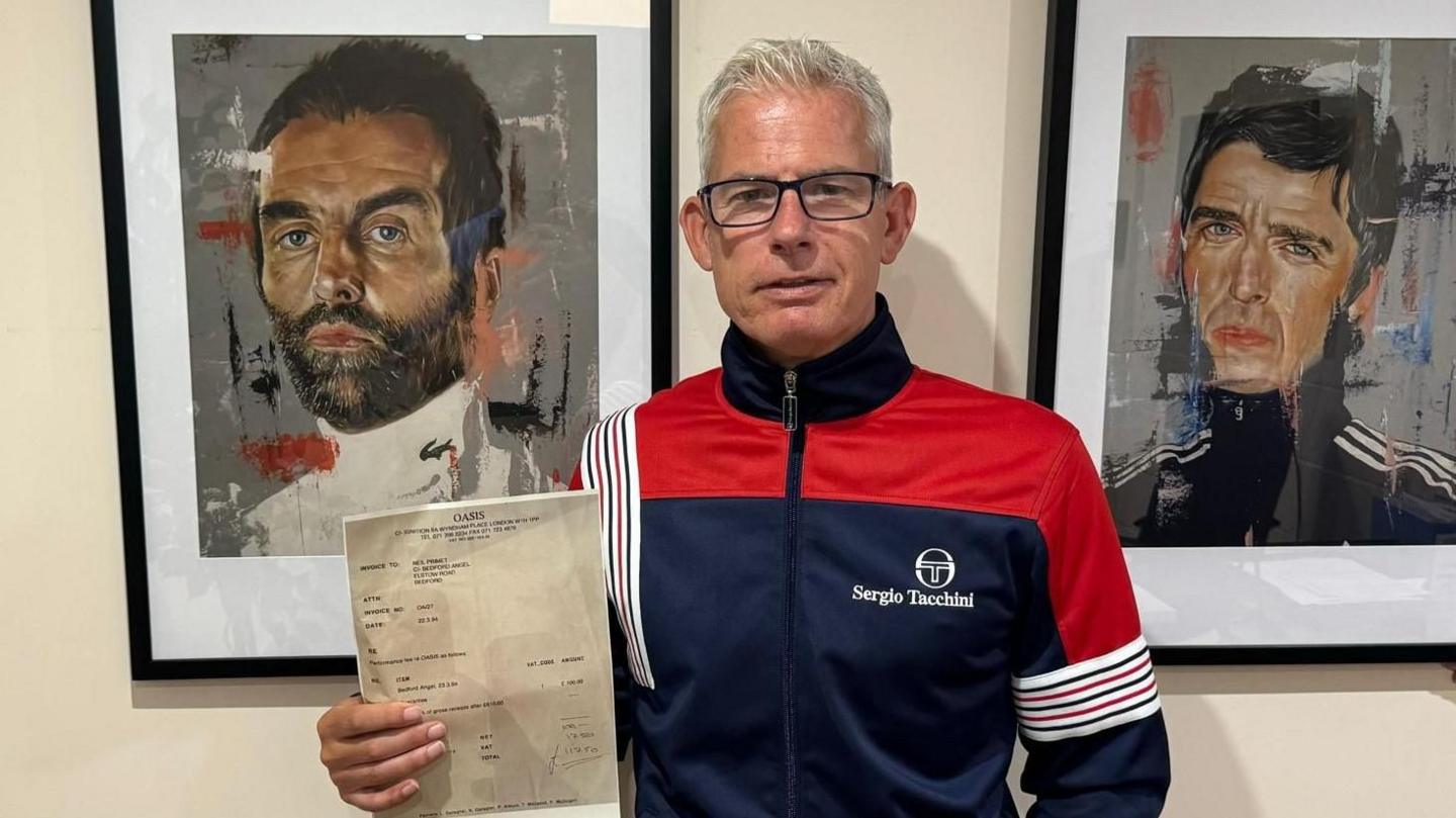 Neil Primett, wearing glasses with grey hair, holding his invoice next to portraits of Noel and Liam Gallagher