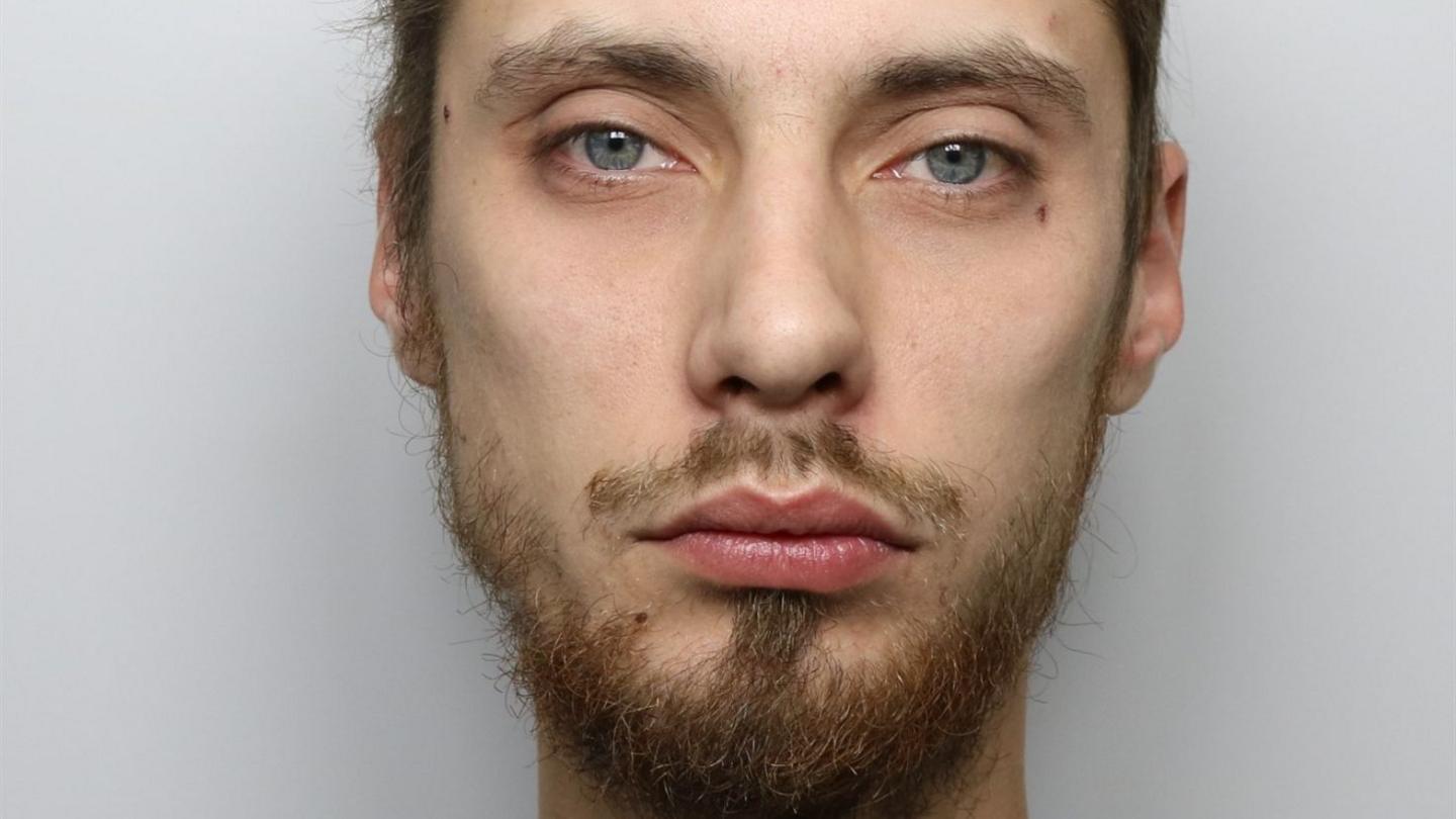 A man with short brown hair and brown or ginger facial hair and blue eyes. It is a custody photo and the background is grey.