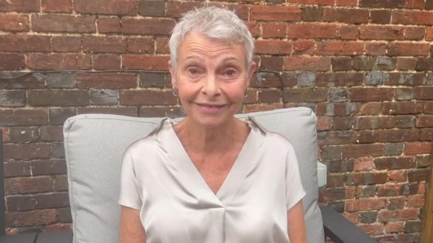 Maggie Hackney pictured outdoors on a garden seat with a brick wall behind her. She smiles at the camera and wears a beige/grey silk short-sleeved shirt with a v-neck and gold hoop earrings. She has short pixie style grey hair.