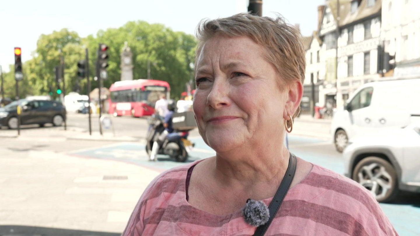 Carole Powell is standing in Clapham High Street on a sunny day. She wears a pink striped top and has short hair and hoop earrings. 