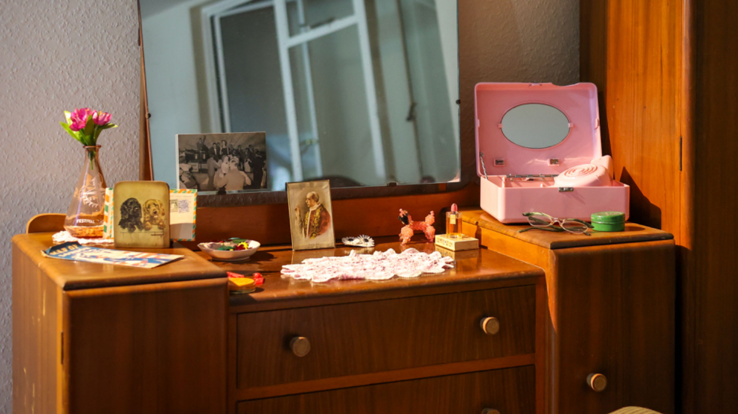 A room set in 1956 which has a dressing table on display that has items including a small vase with a pink flower, old photos, a pair of glasses and a pink make up case